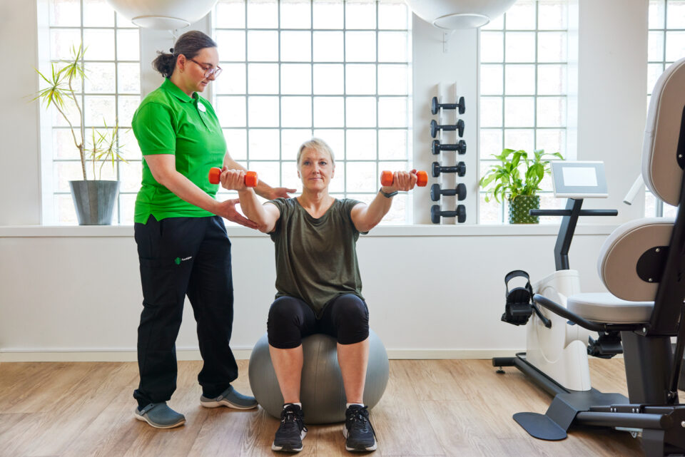 En fysioterapeut visar en patient hur hon ska träna. Patienten sitter på en pilatesboll med en hantel i vardera handen.