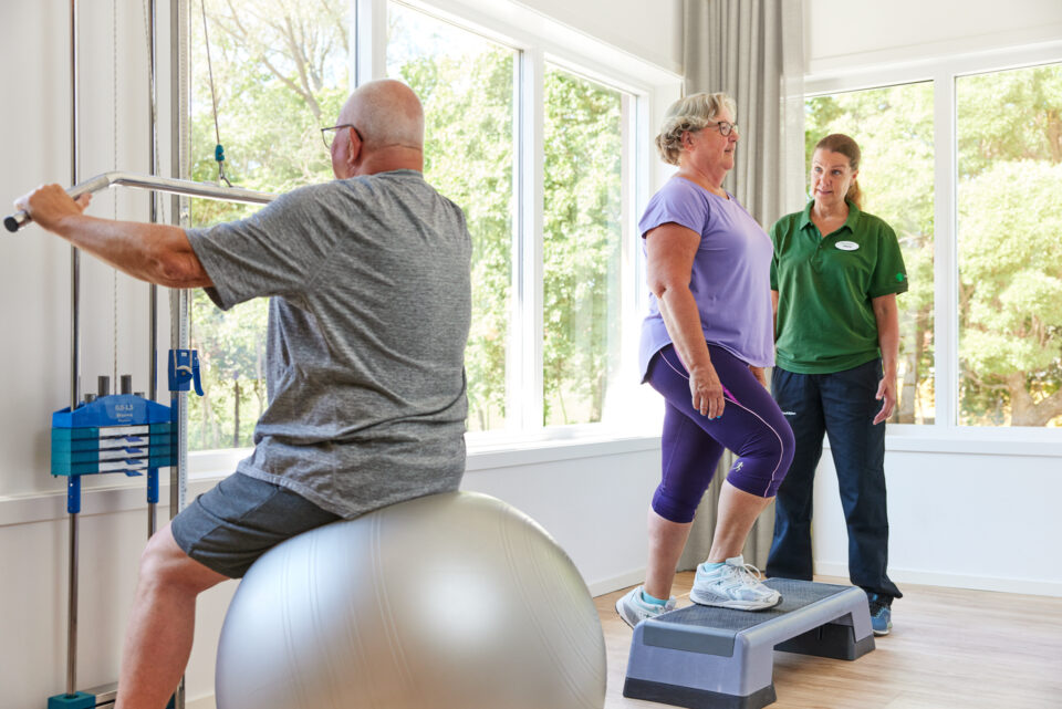 En äldre man sitter på en pilatesboll och tränar armarna i en dragmaskin. En kvinna tränar på en step-upp bräda och en rehabassistent står bredvid och instruerar.