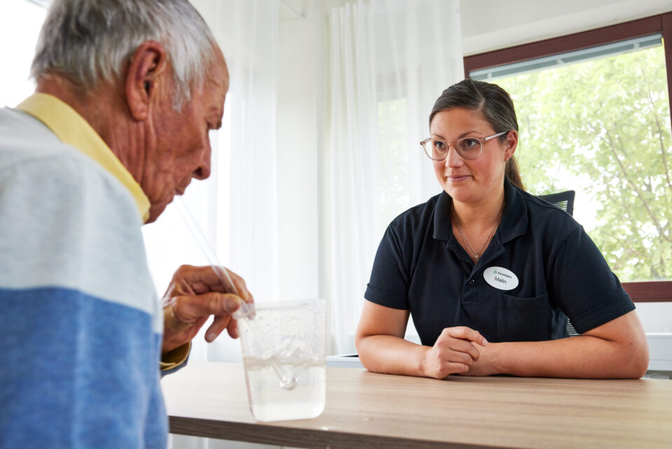 En patient får blåsa med sugrör i ett vattenglas så det bubblar sig. Logopeden sitter mittemot.