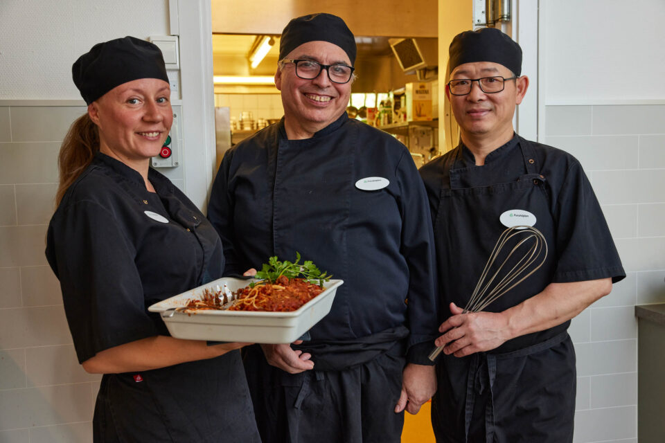 Tre glada kockar poserar. De har svarta arbetskläder på sig. En av kockarna är kvinna och håller i ett serveringsfat med mat.